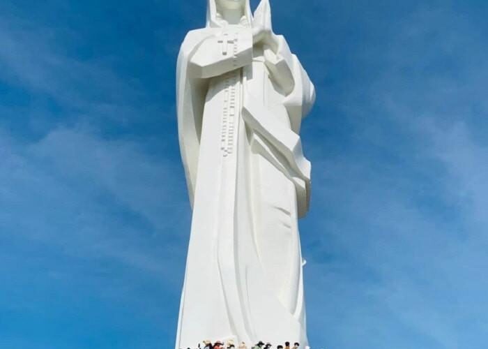 Patung Bunda Maria setinggi tiga meter berwarna putih di puncak Gunung Tà Pao, Bình Thuận, Vietnam, dikelilingi hutan tropis hijau di bawah langit biru cerah.